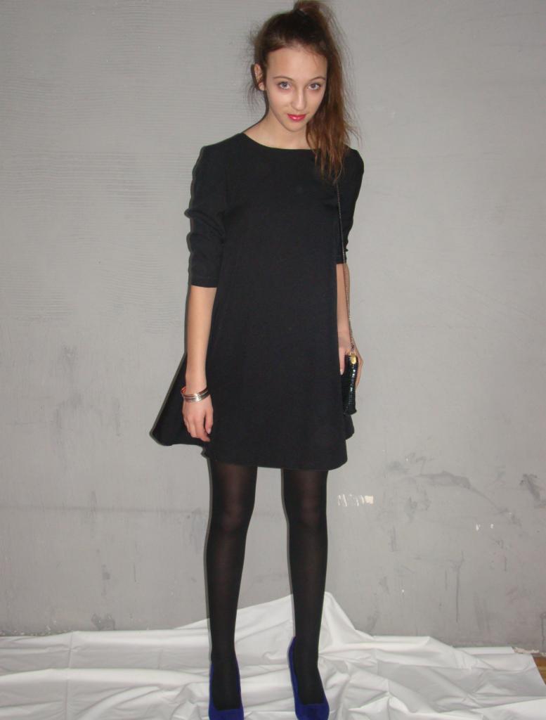 Młoda kobieta w czarnej sukience, czarnych rajstopach i niebieskich butach na obcasie, pozująca na tle szarej ściany i białego materiału.