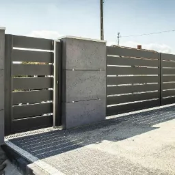 Nowoczesna, grafitowa brama wjazdowa z poziomymi panelami, osadzona w betonowych słupkach, na tle kostki brukowej i błękitnego nieba.