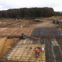 Zbrojenie fundamentów z drewnianym szalunkiem, widoczna siatka z prętów stalowych, piła spalinowa do betonu, w tle las i teren budowy.