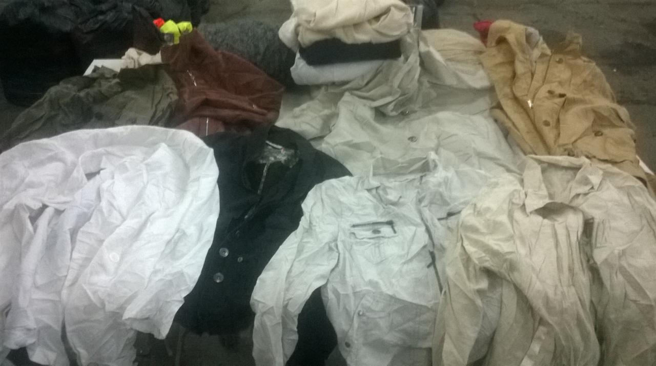 Stos ubrań używanych w różnych kolorach i fasonach, widoczne koszule, marynarki i inne elementy garderoby. Niektóre ubrania są pogniecione.