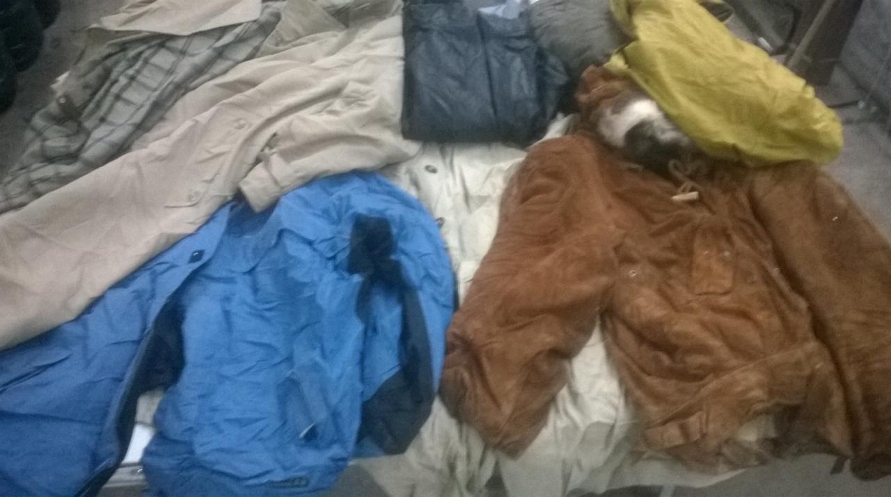 Stos odzieży używanej w różnych kolorach i stylach, w tym niebieska kurtka, brązowa kurtka z kapturem i inne ubrania. Widoczne zagniecenia i różnorodność faktur.