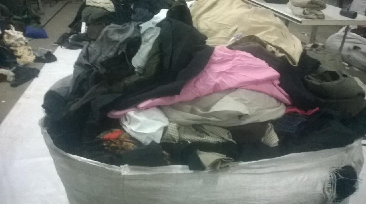 Worek z odzieżą używaną w magazynie. Widoczne ubrania w różnych kolorach i fakturach, częściowo wystające z worka. W tle fragment stołu.