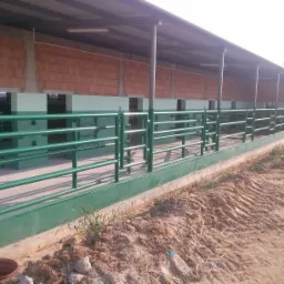 Zewnętrzny widok budynku z zielonymi, metalowymi poręczami. Budynek w trakcie budowy, z odsłoniętymi cegłami i ziemią dookoła. Widoczne oświetlenie pod dachem.