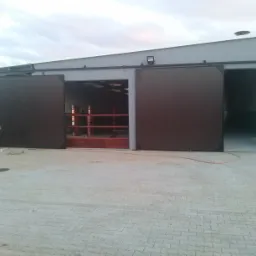 Szara hala z przesuwanymi, brązowymi drzwiami. Widoczny fragment osoby pracującej przy drzwiach oraz wjazd do wnętrza hali.