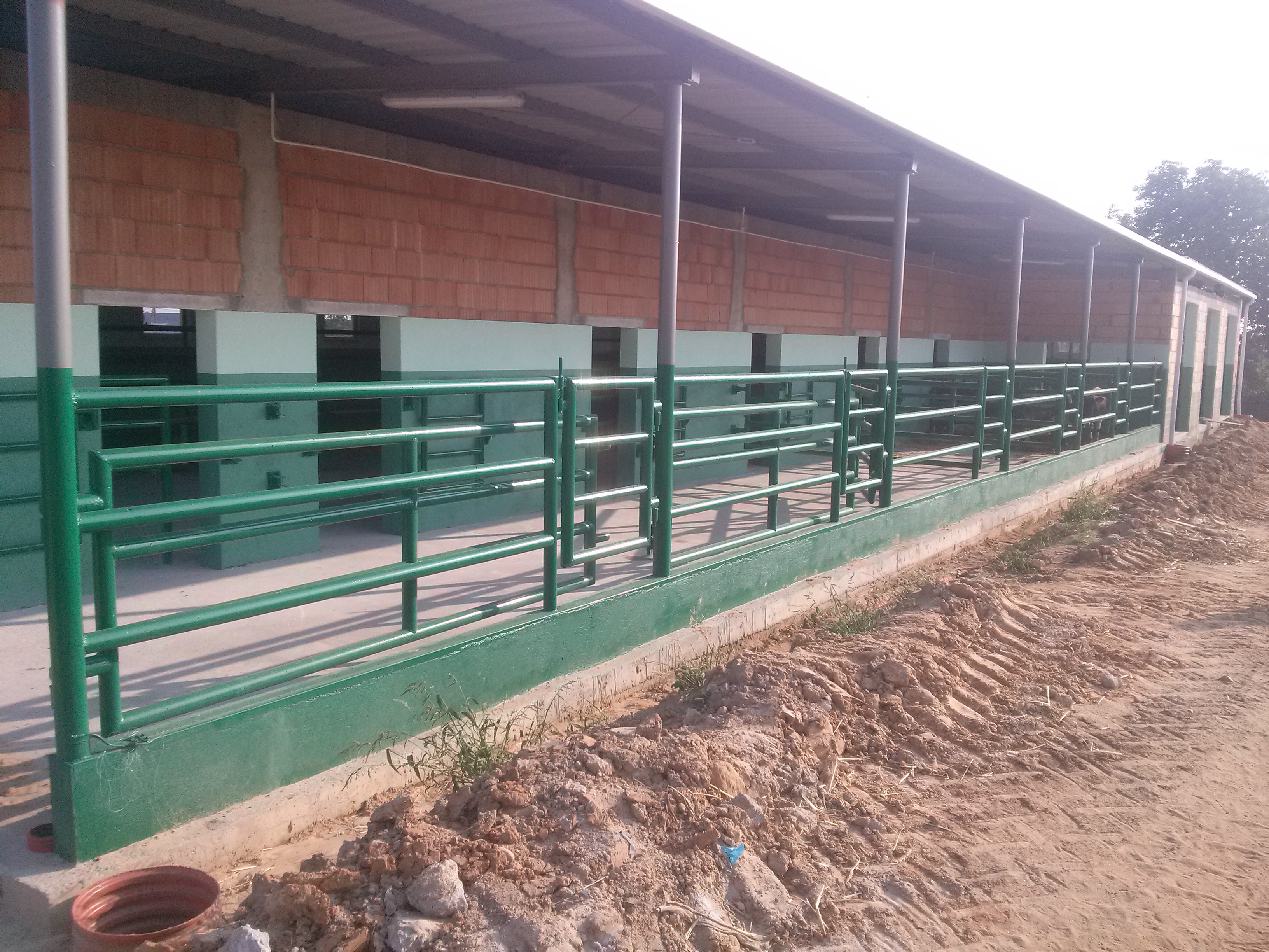 Zewnętrzny widok budynku z zielonymi, metalowymi poręczami. Budynek w trakcie budowy, z odsłoniętymi cegłami i ziemią dookoła. Widoczne oświetlenie pod dachem.