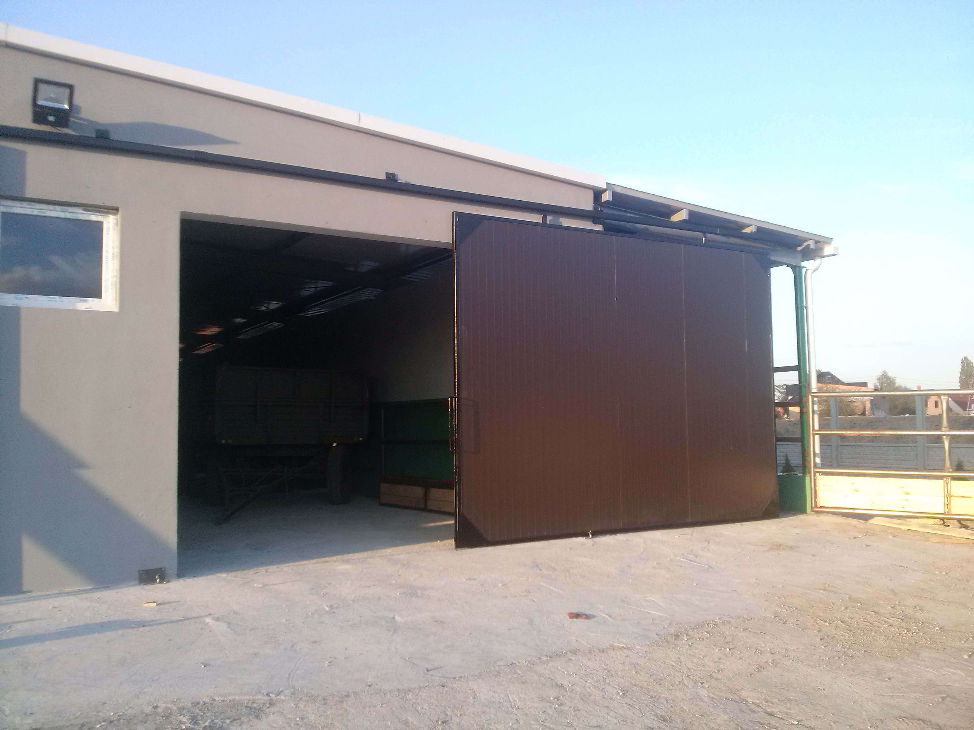 Brązowa, przesuwna brama garażowa z pionowymi panelami, częściowo otwarta, odsłaniająca wnętrze z przyczepą. Budynek z oknem, oświetlenie zewnętrzne.