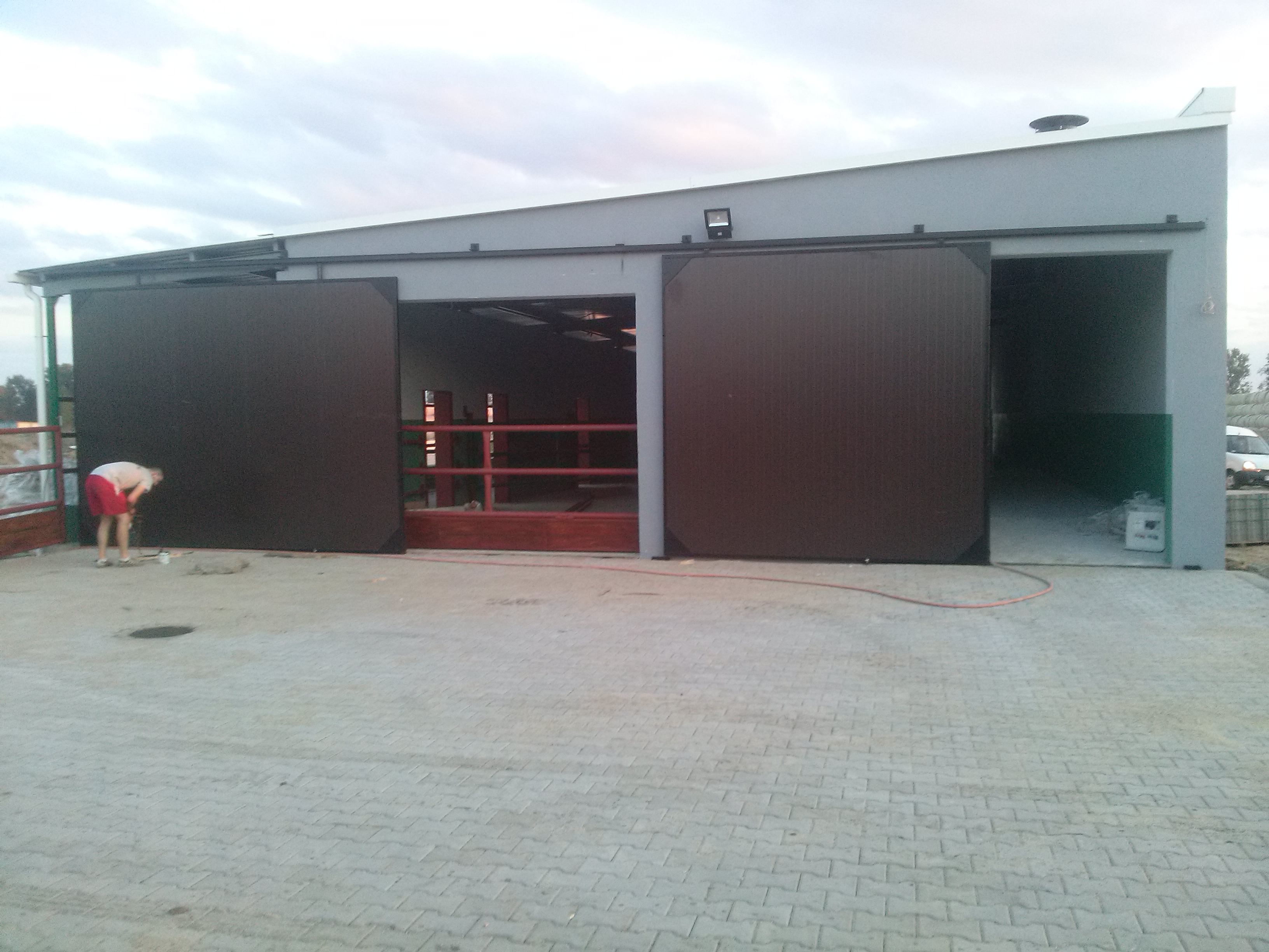 Szara hala z przesuwanymi, brązowymi drzwiami. Widoczny fragment osoby pracującej przy drzwiach oraz wjazd do wnętrza hali.