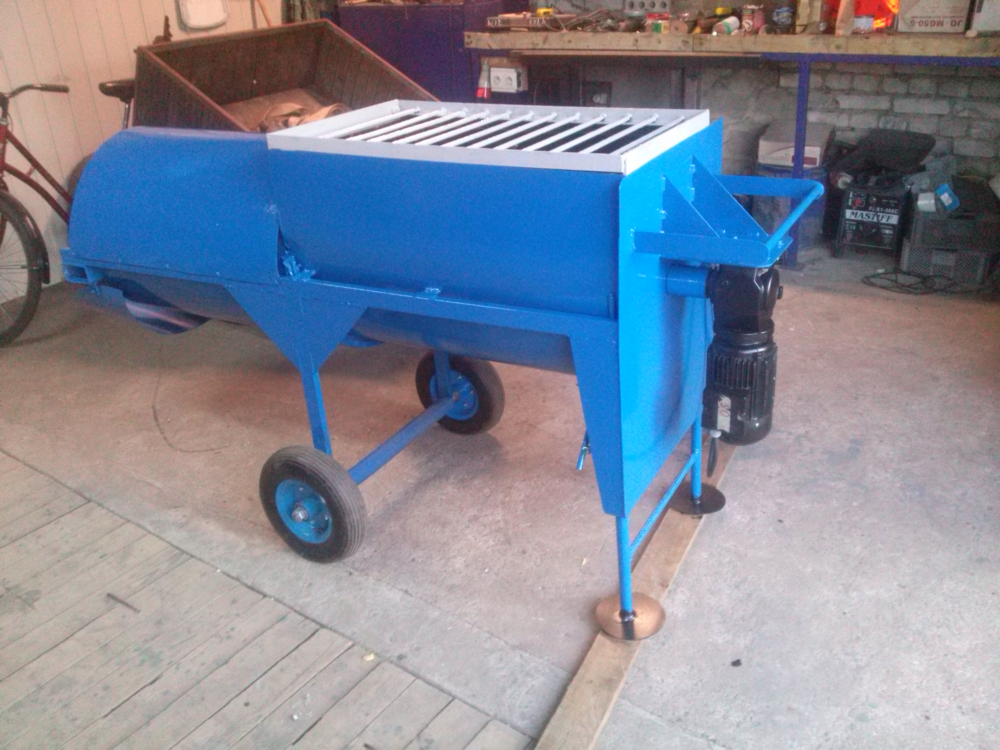 Nietypowa, niebieska konstrukcja metalowa na kółkach z silnikiem elektrycznym, przypominająca maszynę rolniczą lub przemysłową, stoi w warsztacie.