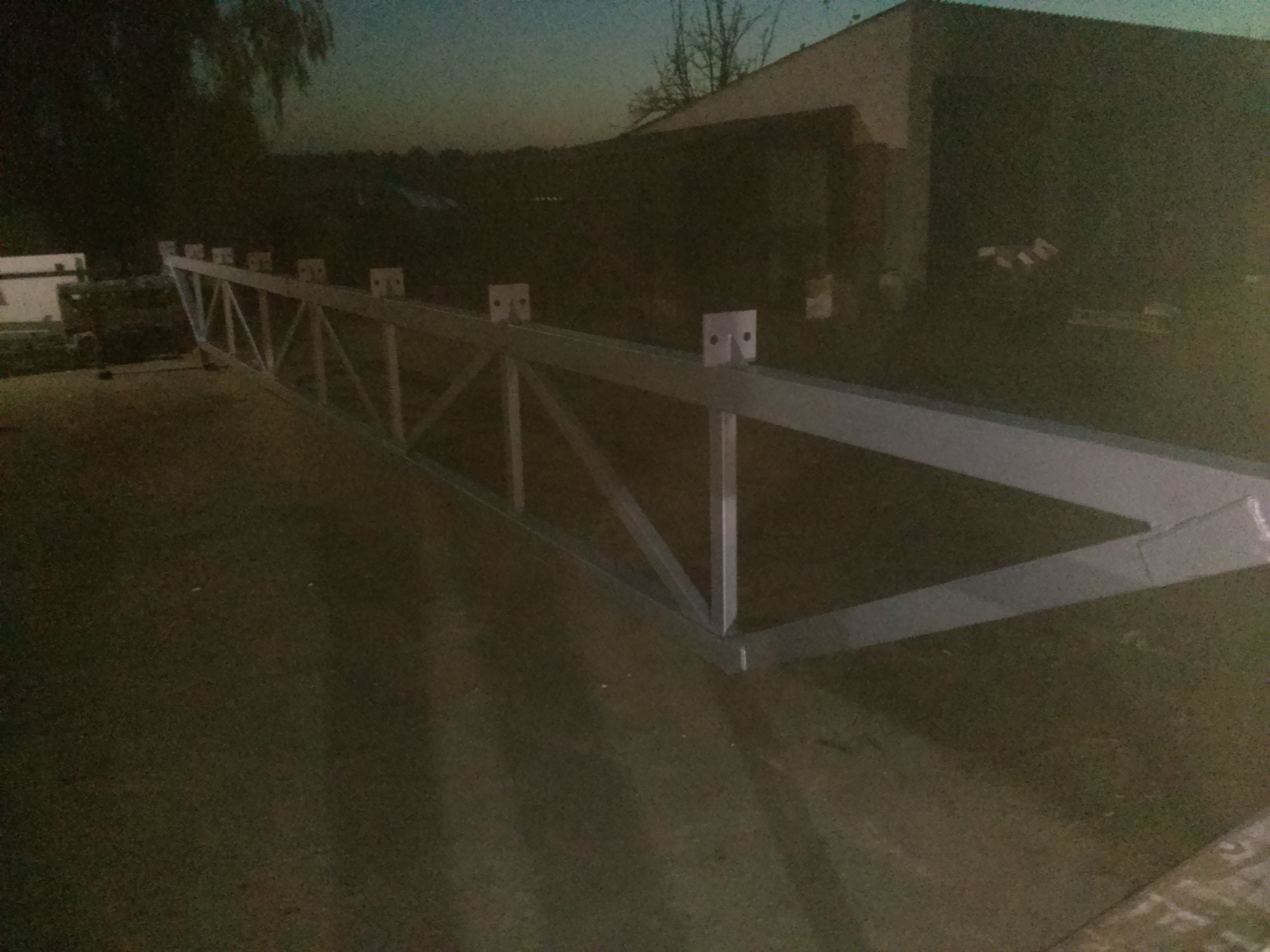 Metalowa konstrukcja pergoli z widocznymi elementami montażowymi, leżąca na ziemi przed budynkiem w ciemnym otoczeniu. Elementy spawane, gotowe do montażu.