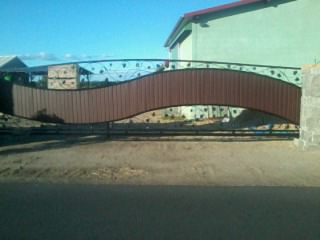 Falista brama wjazdowa z brązowym wypełnieniem i ozdobnymi elementami roślinnymi, na tle błękitnego nieba i budynku.