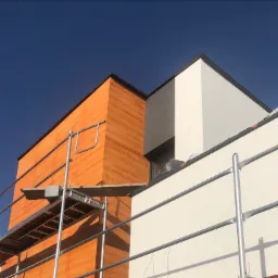 Fragment elewacji budynku z pomarańczową okładziną drewnopodobną i białym fragmentem, widoczne rusztowanie z deską, błękitne niebo w tle.