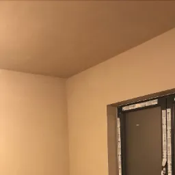 Świeżo otynkowane ściany i sufit w pomieszczeniu z oknem zabezpieczonym taśmą malarską, gotowe do dalszych prac wykończeniowych.