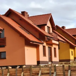Szereg nowych domów z czerwonymi dachami w pastelowych kolorach, widoczne zza drewnianego ogrodzenia na nierównym terenie.