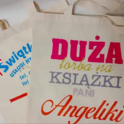 Dwie beżowe torby bawełniane z kolorowymi nadrukami: jedna z logo firmy 'Świątl', druga z napisem 'DUŻA torba na KSIĄŻKI PANI Angeliki' w różnych kolorach.