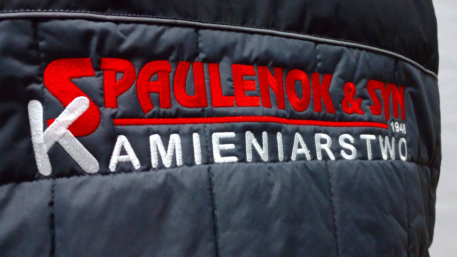 Fragment pikowanej, ciemnej kurtki roboczej z czerwono-białym haftowanym logo firmy 'SPAULENOK & SYN KAMIENIARSTWO 1948'.