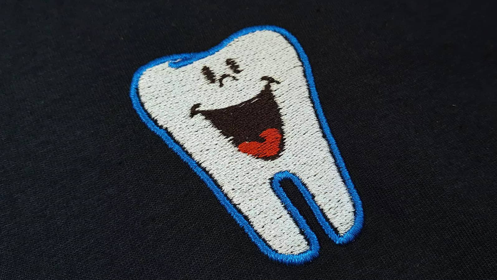 Czarna koszulka z haftowanym, kreskówkowym zębem z niebieską obwódką, uśmiechniętą buzią i czerwonym językiem.