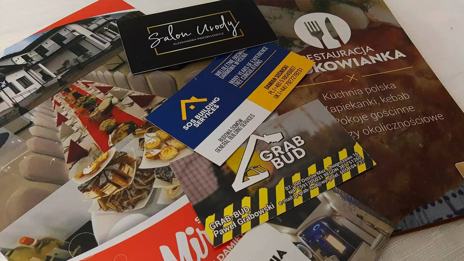 Stos wizytówek i ulotek reklamowych różnych firm, w tym salonu urody, firmy budowlanej i restauracji, ułożonych warstwowo na białym tle.