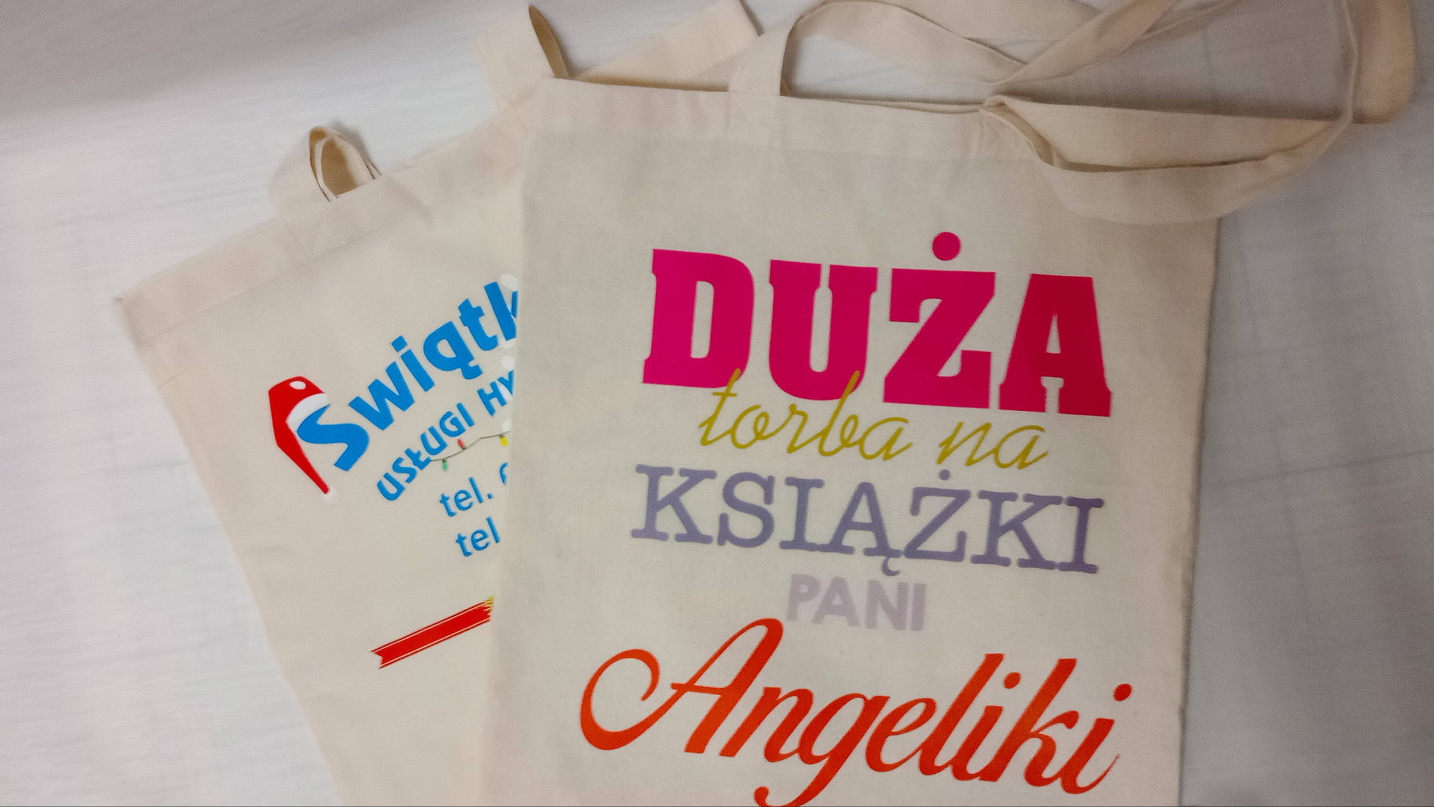Dwie beżowe torby bawełniane z kolorowymi nadrukami: jedna z logo firmy 'Świątl', druga z napisem 'DUŻA torba na KSIĄŻKI PANI Angeliki' w różnych kolorach.