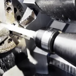 Precyzyjna obróbka metalu na tokarce CNC. Fragment białego pręta w uchwycie, skrawanie narzędziem. Detal z widocznymi śladami chłodziwa.