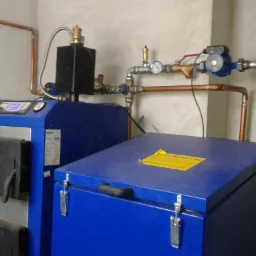 Niebieski piec CO z czarnymi drzwiczkami i instalacją miedzianą, widoczny w kotłowni. Na górze pieca żółta naklejka informacyjna.