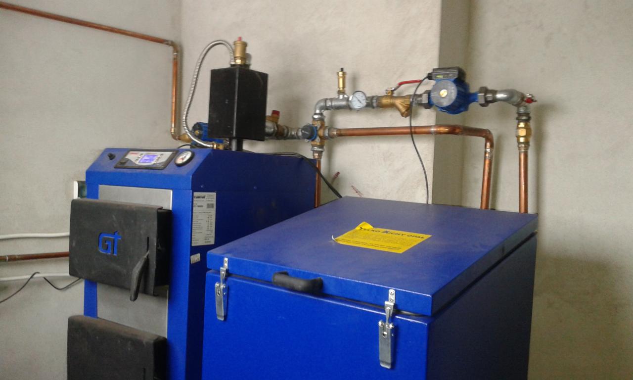 Niebieski piec CO z czarnymi drzwiczkami i instalacją miedzianą, widoczny w kotłowni. Na górze pieca żółta naklejka informacyjna.