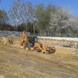 Żółta koparko-ładowarka CASE na placu budowy, przygotowująca teren pod fundamenty, widoczne stalowe zbrojenia, w tle drzewa i ogrodzenie.