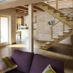 Wnętrze domu z bali: drewniane schody ze stalowymi poręczami, widok z salonu z fioletową sofą na otwartą kuchnię, akwarium i dekoracja ścienna w kształcie ptaka.