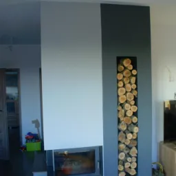 Nowoczesny kominek z wnęką na drewno, pomalowany w odcieniach szarości, z widocznym paleniskiem i ułożonymi szczapami drewna w dekoracyjnej wnęce.