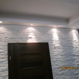 Ściana wykończona białym kamieniem dekoracyjnym z ciemnymi drzwiami w nowoczesnym wnętrzu, oświetlona punktowymi lampami sufitowymi, z widocznym fragmentem sufitu.