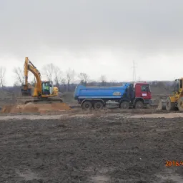 Żółta koparka, niebiesko-czerwona wywrotka i żółta ładowarka na placu budowy w pochmurny dzień. Koparka nabiera ziemię, a na wywrotce widoczny napis TRANS-TATRA.
