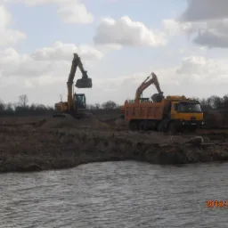 Dwie żółte koparki i wywrotka Tatra podczas prac ziemnych na brzegu zbiornika wodnego, widoczne ślady opon na błotnistym terenie, pochmurne niebo.