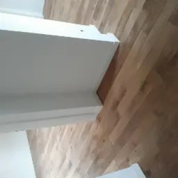 Ukośne ujęcie świeżo położonej, jasnobrązowej podłogi z drewnianych desek, widoczna biała listwa przypodłogowa.