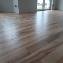 Jasna, nowa podłoga z paneli drewnopodobnych ułożona w pustym, jasnym pomieszczeniu z dużym oknem balkonowym.