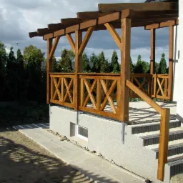 Drewniana konstrukcja z balustradą i schodami przy budynku, z widocznymi elementami zadaszenia i otaczającą zielenią. Ujęcie z dołu, podkreślające solidność wykonania.