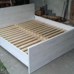 Białe łóżko z drewnianym stelażem, widoczne deski i elementy konstrukcyjne. Surowe tło warsztatowe. Kątowe ujęcie, podkreślające prostotę formy.