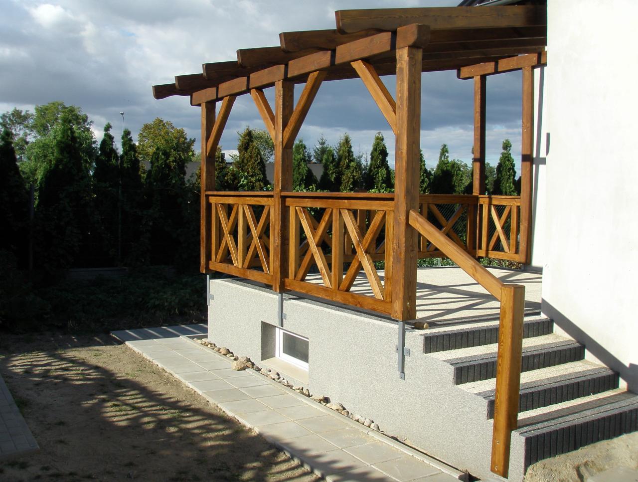 Drewniana konstrukcja z balustradą i schodami przy budynku, z widocznymi elementami zadaszenia i otaczającą zielenią. Ujęcie z dołu, podkreślające solidność wykonania.