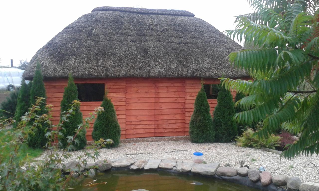 Oryginalny domek ogrodowy z drewnianą elewacją i strzechą, otoczony zielenią i oczkiem wodnym. Nietypowe pokrycie dachowe.