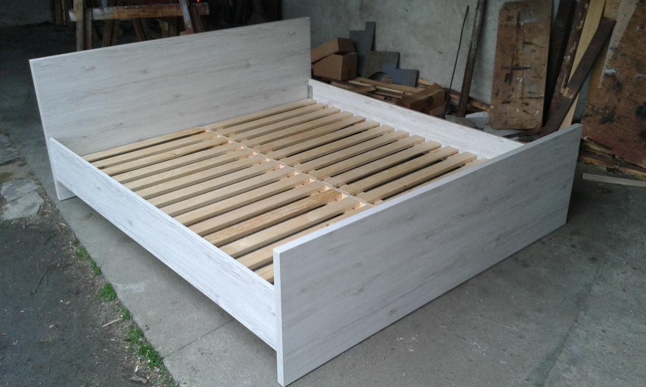 Białe łóżko z drewnianym stelażem, widoczne deski i elementy konstrukcyjne. Surowe tło warsztatowe. Kątowe ujęcie, podkreślające prostotę formy.