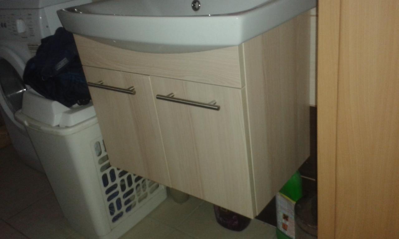 Jasna, wisząca szafka pod umywalkę z dwoma drzwiczkami i uchwytami, w tle pralka i kosz na pranie. Ujęcie z bliska, w łazience.