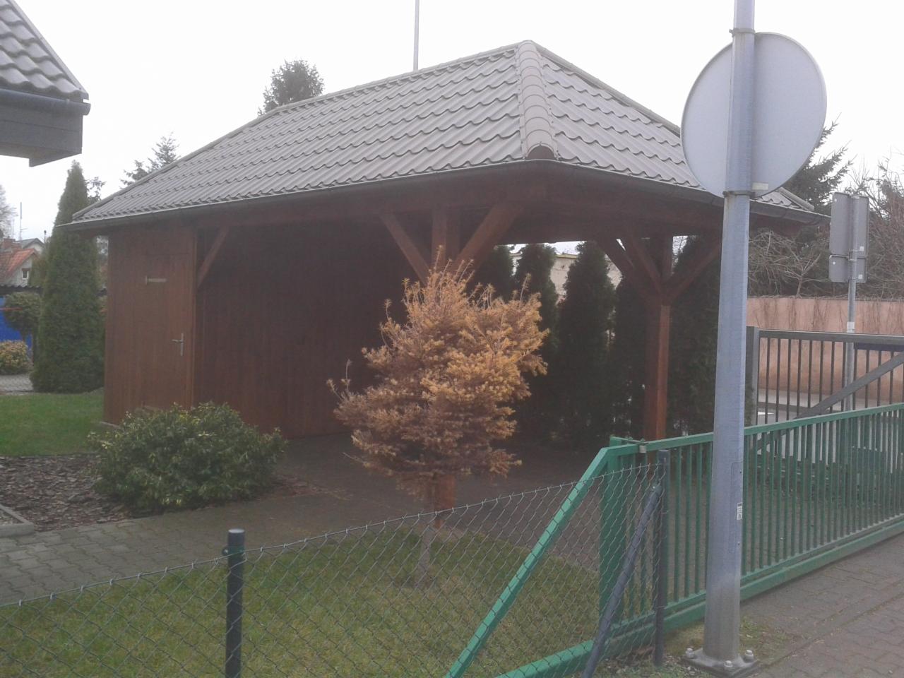 Zadaszona, drewniana konstrukcja z metalowym dachem, widoczna zza zielonego ogrodzenia. Na pierwszym planie uschnięte drzewko, w tle iglaki i budynek.