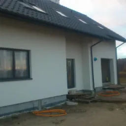 Niedokończony biały dom z czarną dachówką i oknami, widoczne fundamenty i teren wokół budynku w trakcie prac wykończeniowych, pomarańczowe rury leżące na ziemi.