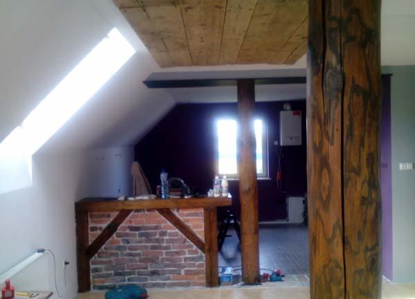 Poddasze z oknem dachowym i drewnianym wykończeniem sufitu, ceglanym barem oraz grubą, drewnianą kolumną. Widoczny fragment pokoju z oknem.