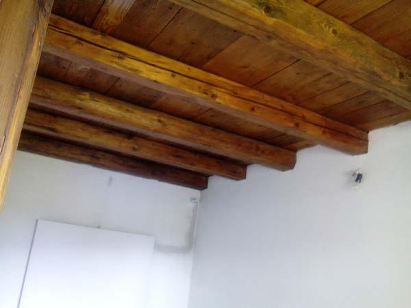 Rustykalny strop z drewnianych belek w odcieniach brązu, kontrastujący z białą ścianą i widocznym fragmentem instalacji elektrycznej.