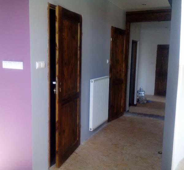 Wnętrze z dwiema otwartymi, drewnianymi drzwiami, biały grzejnik na szarej ścianie, fioletowy fragment ściany po lewej. Podłoga surowa, w tle widoczne kolejne drzwi.