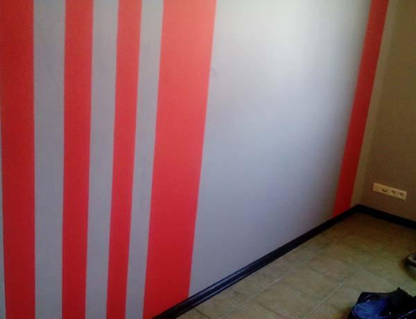 Ściana w pionowe, czerwono-szare pasy, świeżo pomalowana, widoczna podłoga z listwami przypodłogowymi i gniazdko elektryczne. Efekt tapetowania lub malowania dekoracyjnego.