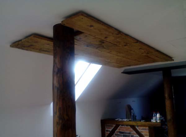Rustykalny detal wnętrza: drewniany słup i belki stropowe z widocznymi słojami, oświetlone naturalnym światłem z okna dachowego. Tło stanowi jasna ściana.