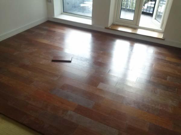 Błyszcząca, brązowa podłoga z parkietu ułożona w jasnym pokoju z dużym oknem. Na podłodze leży pojedyncza deska parkietowa.