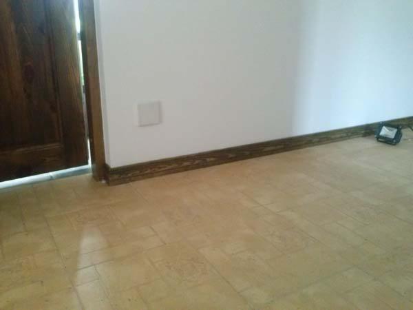 Jasna podłoga z paneli, drewniana listwa przypodłogowa wzdłuż białej ściany, fragment drewnianych drzwi po lewej. Widoczny kontakt i lampa podłogowa.