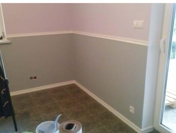 Pomieszczenie po malowaniu ścian na szaro i różowo, z widocznymi listwami przypodłogowymi i narzędziami malarskimi w rogu. Widoczny fragment okna balkonowego.