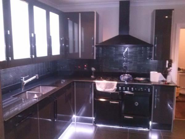 Nowoczesna kuchnia w ciemnej tonacji z błyszczącymi frontami, podświetleniem LED i czarnym okapem. Widoczne okna i armatura. Minimalistyczny design.